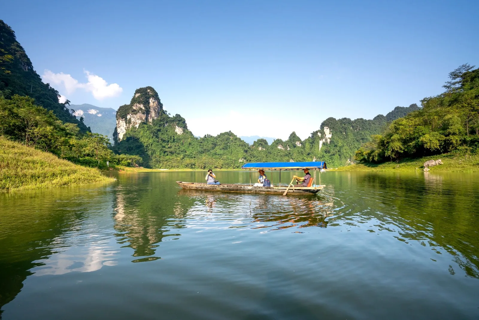 A boat with a blue canopy carries travelers past limestone peaks on a calm river in Laos.